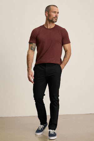 Man with short hair and a trimmed beard wears the MADDOX PANT by Velvet by Graham & Spencer, paired with a maroon t-shirt and navy sneakers, standing against a plain light background for a polished look.