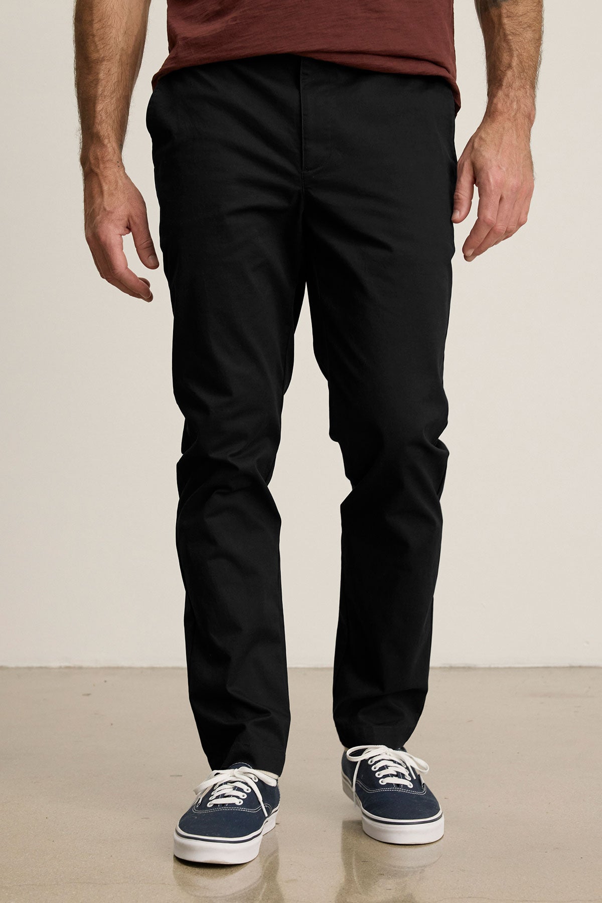 Someone stands on a smooth beige floor against a plain light background, wearing Velvet by Graham & Spencer’s MADDOX PANT in black stretch cotton twill, paired with a maroon shirt and navy blue sneakers.-39599110684865