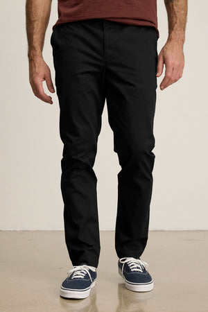 Someone stands on a smooth beige floor against a plain light background, wearing Velvet by Graham & Spencer’s MADDOX PANT in black stretch cotton twill, paired with a maroon shirt and navy blue sneakers.