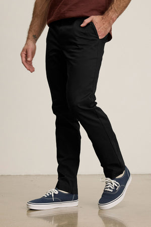 Wearing Velvet by Graham & Spencer’s MADDOX PANT in stretch cotton twill, tailored black style, paired with blue sneakers and a maroon shirt, the person stands with one hand in their pocket on a polished floor for a refined look.