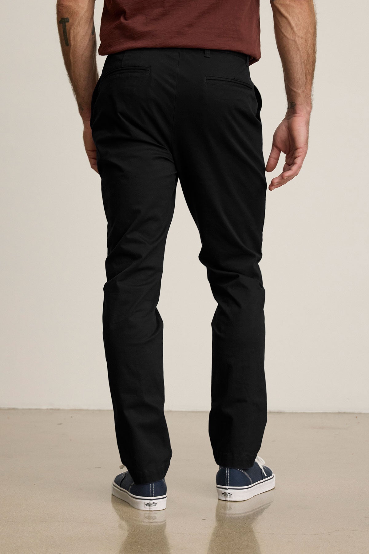 A man stands facing away on a light floor, wearing the MADDOX PANT by Velvet by Graham & Spencer in tailored black stretch cotton twill, paired with a maroon shirt and blue sneakers.-39599110750401