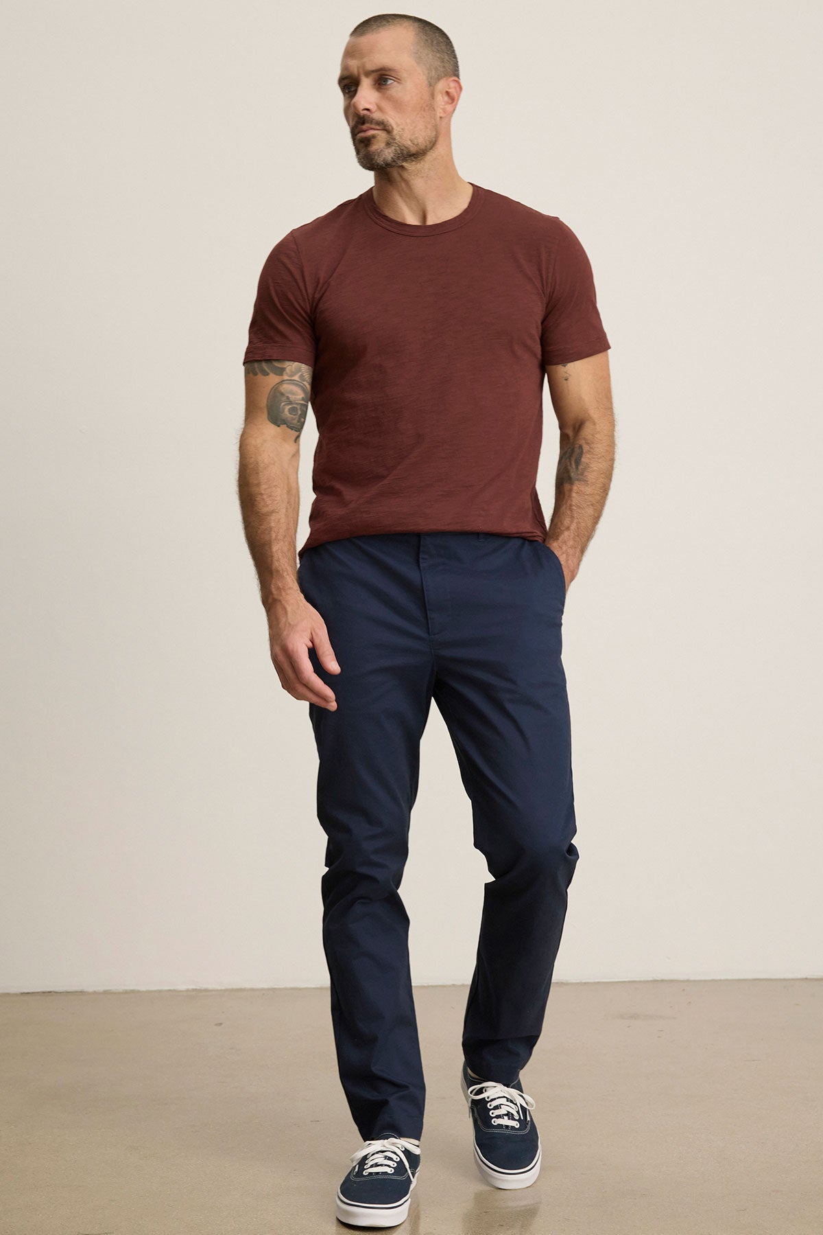Man stands against a plain background, hands in pockets and a neutral expression, wearing a maroon t-shirt, dark sneakers, and Velvet by Graham & Spencer's MADDOX PANT in navy blue stretch cotton twill.