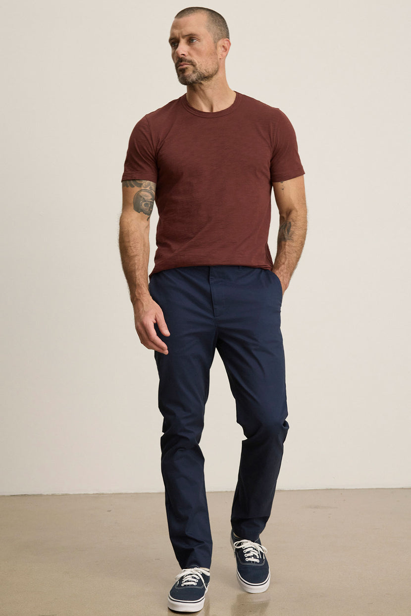 Man stands against a plain background, hands in pockets and a neutral expression, wearing a maroon t-shirt, dark sneakers, and Velvet by Graham & Spencer's MADDOX PANT in navy blue stretch cotton twill.