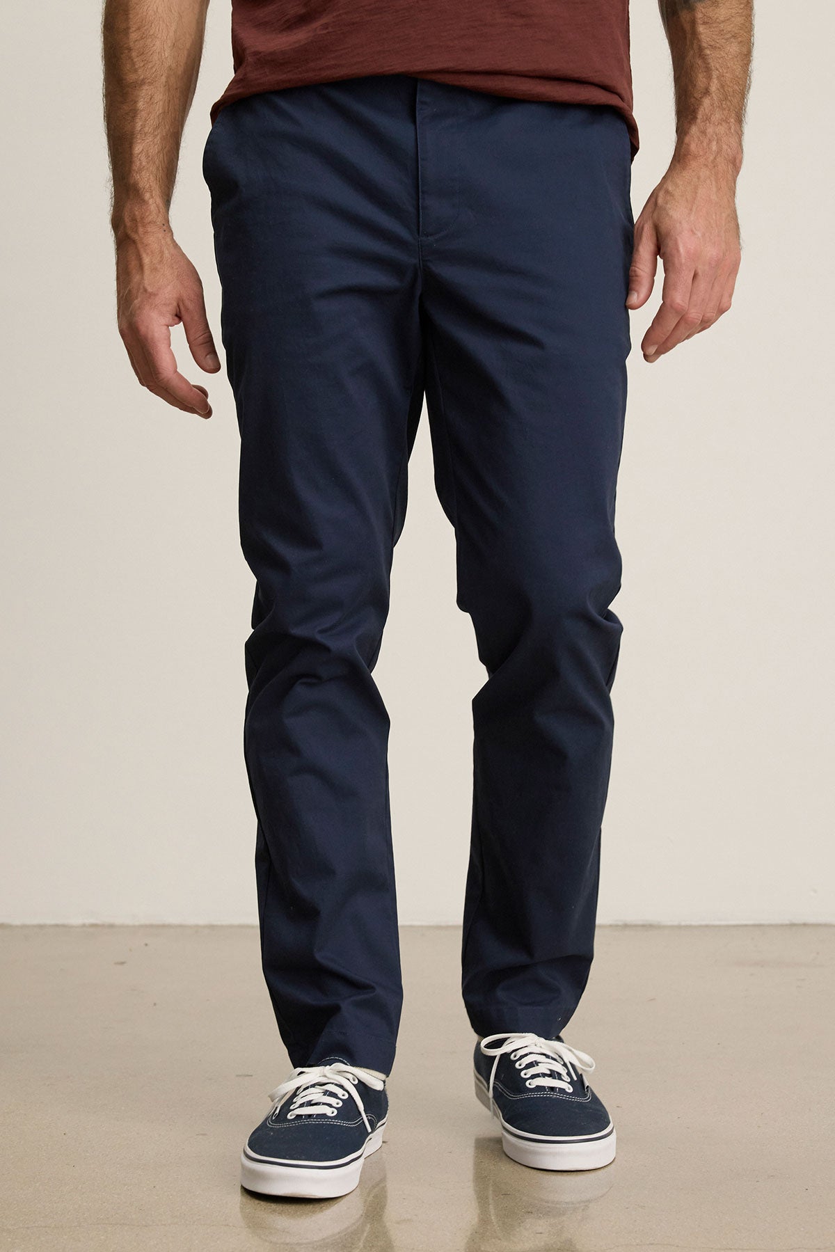 A person stands on a light-colored floor in a maroon shirt, Velvet by Graham & Spencer’s tailored navy MADDOX PANT, and navy sneakers with white soles, creating a polished look.