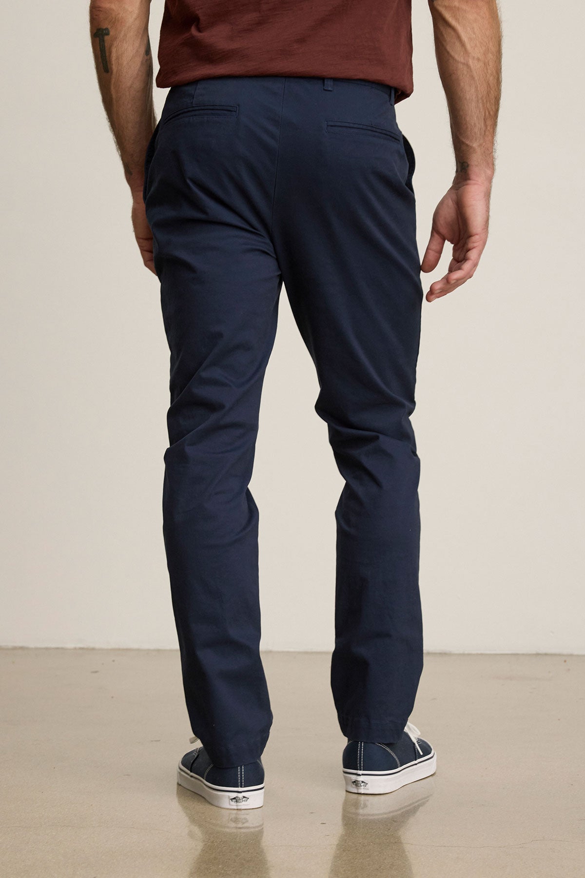 Man stands with his back to the camera, wearing the MADDOX PANT by Velvet by Graham & Spencer in stretch cotton twill, paired with a brown shirt and white sneakers on a light-colored floor.