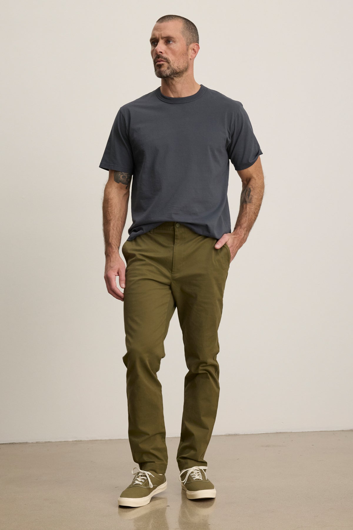 A man stands indoors against a plain light-colored wall, wearing a dark gray t-shirt, Velvet by Graham & Spencer MADDOX PANT in olive green stretch cotton twill, and beige sneakers.-39680477823169