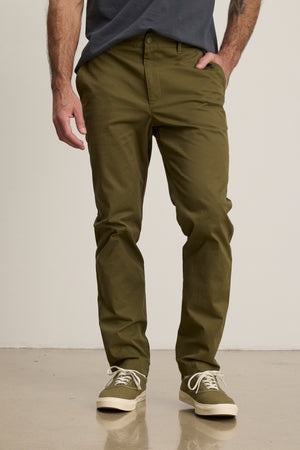 A person stands against a plain light background wearing the MADDOX PANT in olive green by Velvet by Graham & Spencer, paired with a dark t-shirt and matching sneakers.