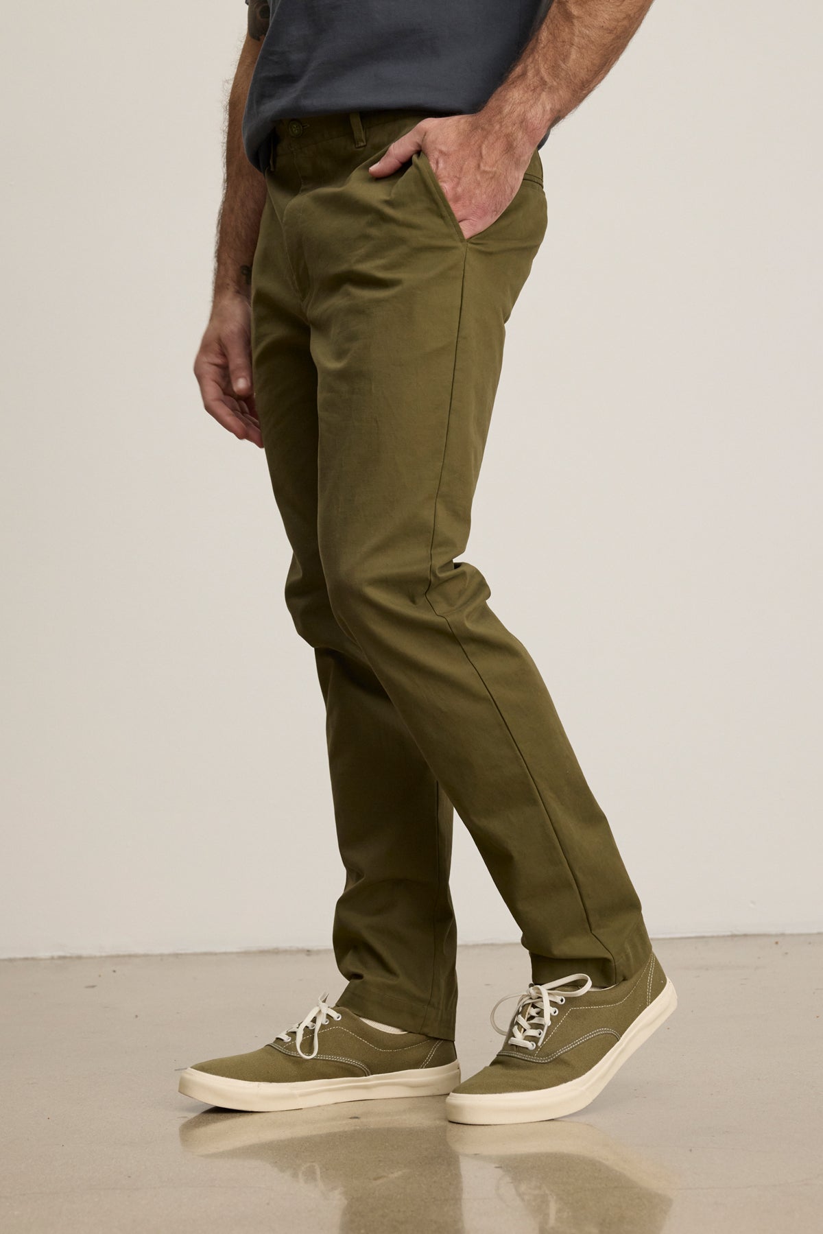 A person in Velvet by Graham & Spencer’s MADDOX PANT—tailored olive green stretch cotton twill—paired with a dark shirt and olive sneakers stands indoors with their left hand in their pocket, creating a refined, polished look.-39680477692097