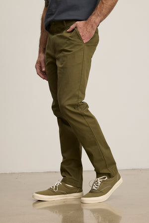 A person in Velvet by Graham & Spencer’s MADDOX PANT—tailored olive green stretch cotton twill—paired with a dark shirt and olive sneakers stands indoors with their left hand in their pocket, creating a refined, polished look.