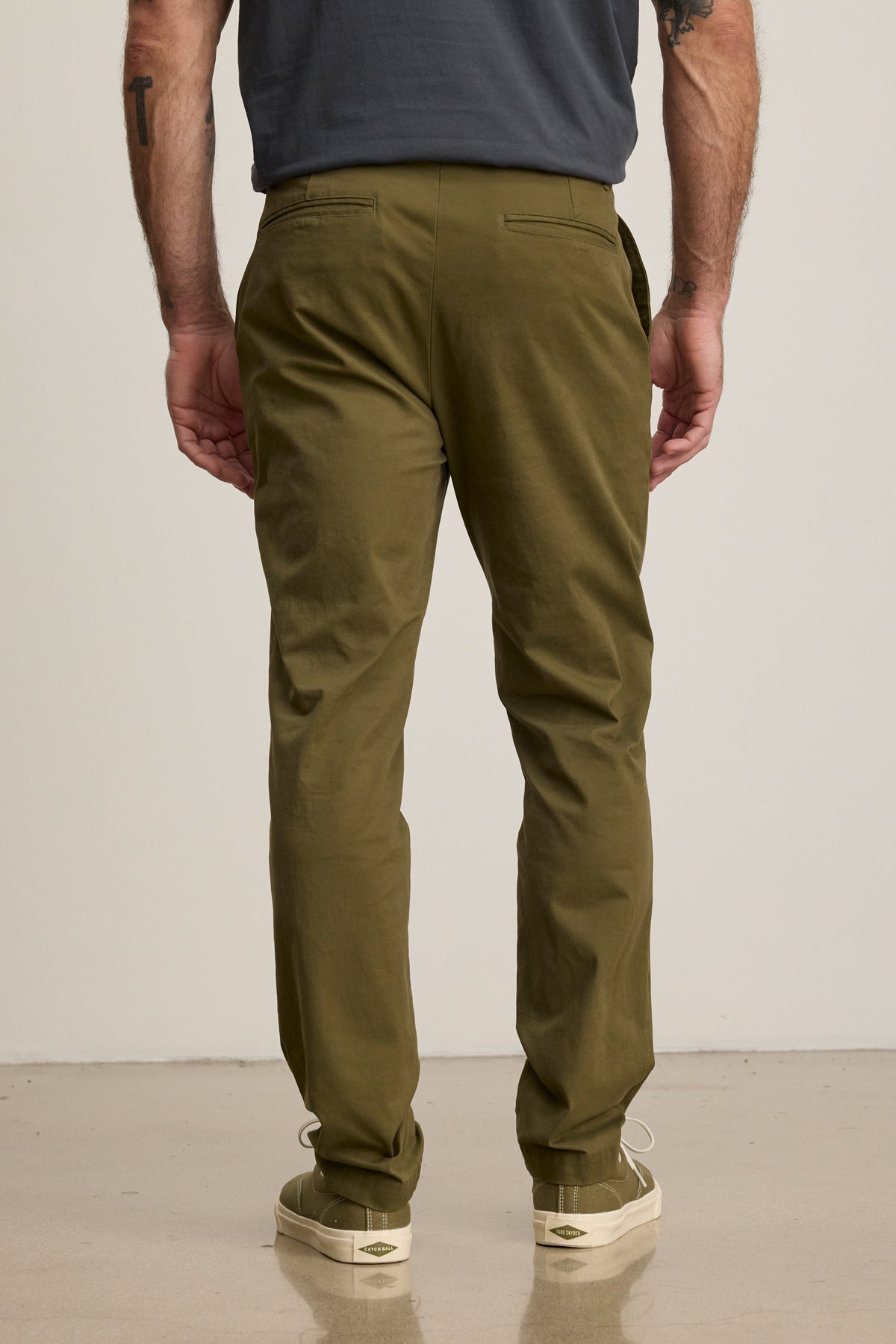 A man stands facing away from the camera in Velvet by Graham & Spencer's MADDOX PANT, tailored in olive green stretch cotton twill, paired with a dark t-shirt and off-white sneakers against a plain background.-39680477659329