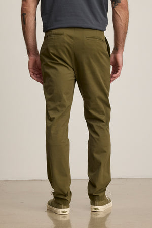 A man stands facing away from the camera in Velvet by Graham & Spencer's MADDOX PANT, tailored in olive green stretch cotton twill, paired with a dark t-shirt and off-white sneakers against a plain background.