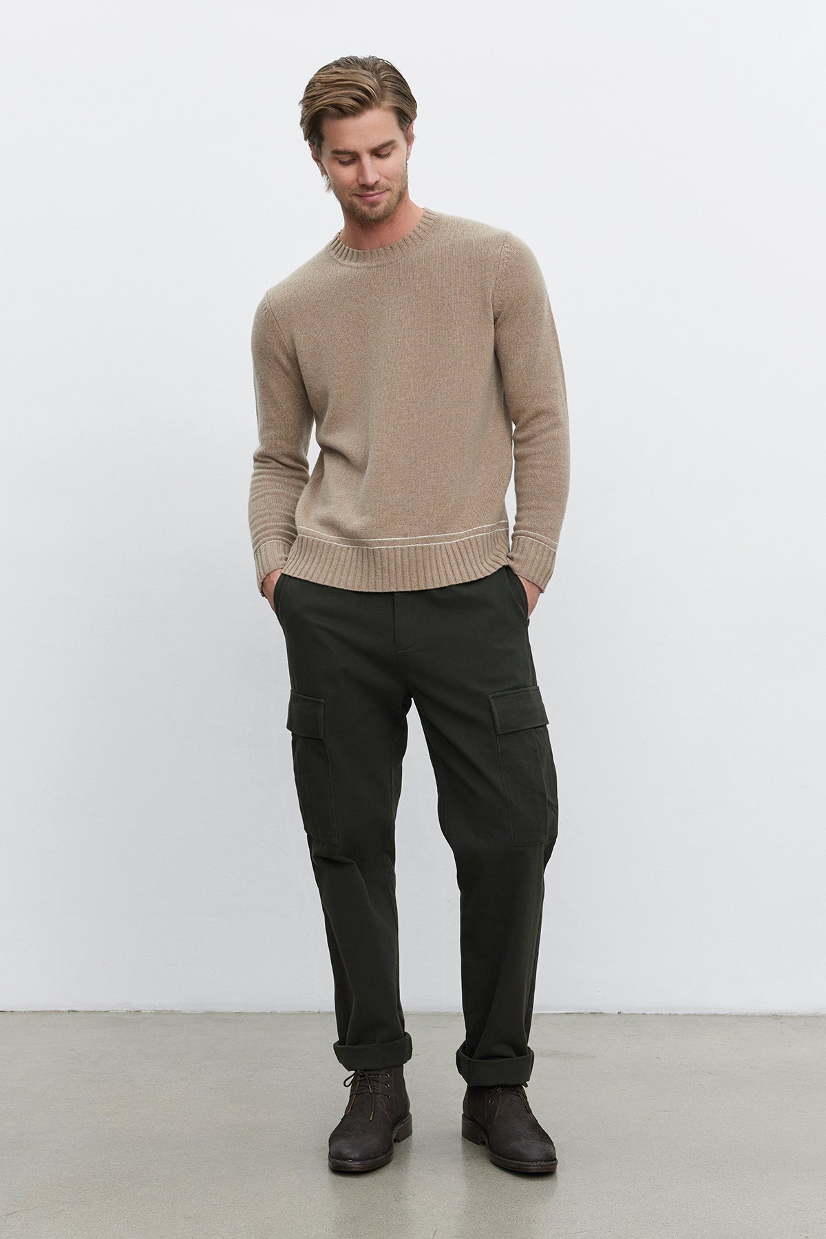   A man with light brown hair wears a beige sweater, the Bryce Pants from Velvet by Graham & Spencer, which are dark green straight-leg pants made of mid-weight cotton lyocell twill, and dark brown shoes. He stands with his hands in his pockets against a plain white background. 