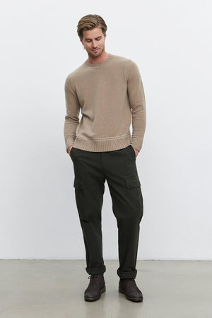 A man with light brown hair wears a beige sweater, the Bryce Pants from Velvet by Graham & Spencer, which are dark green straight-leg pants made of mid-weight cotton lyocell twill, and dark brown shoes. He stands with his hands in his pockets against a plain white background.