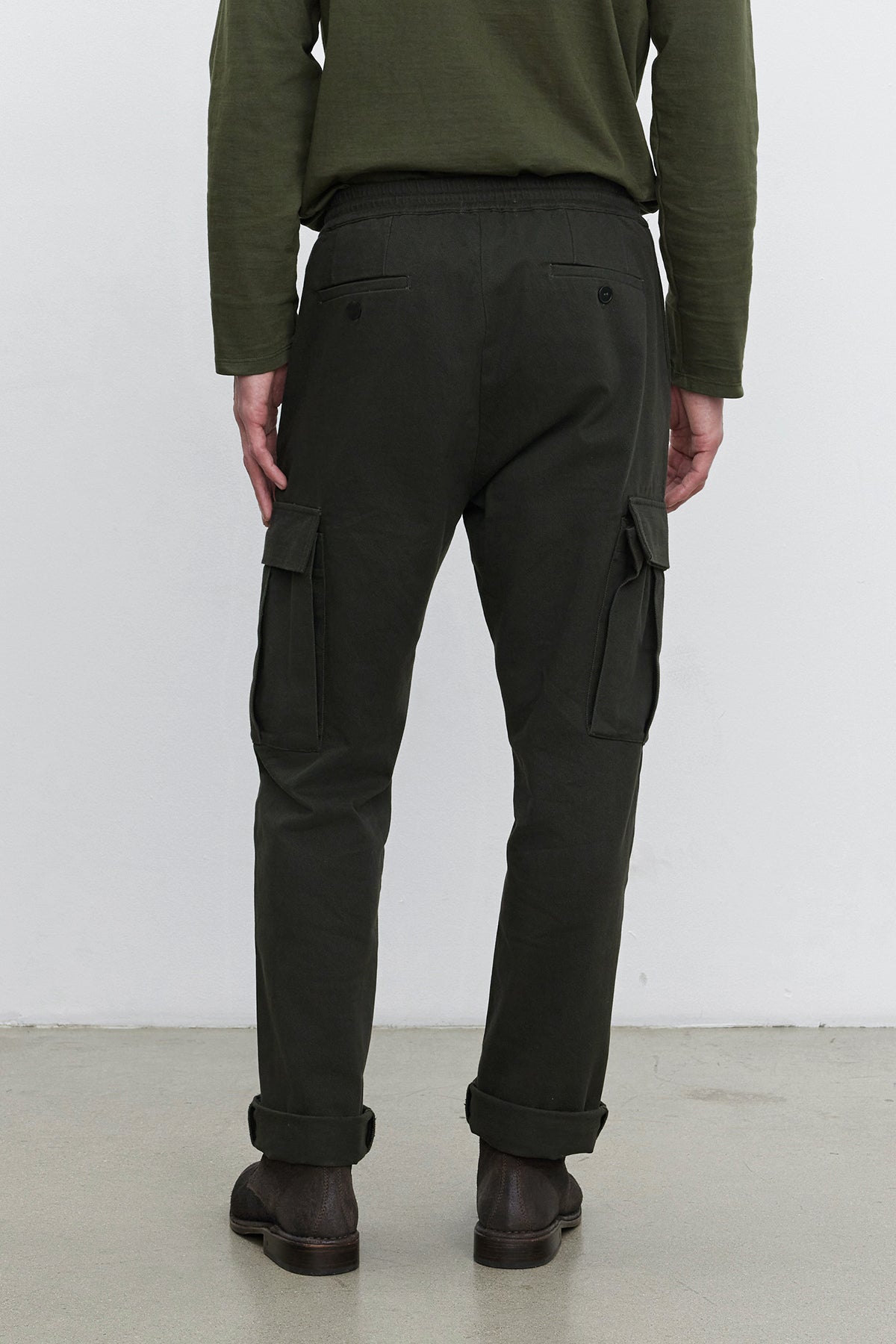   A person stands facing away, wearing a dark green long-sleeve shirt and the BRYCE PANT from Velvet by Graham & Spencer, which are straight leg pants made of dark green cotton lyocell twill with cargo pockets. They are also wearing brown shoes, with the pant legs slightly rolled up. 