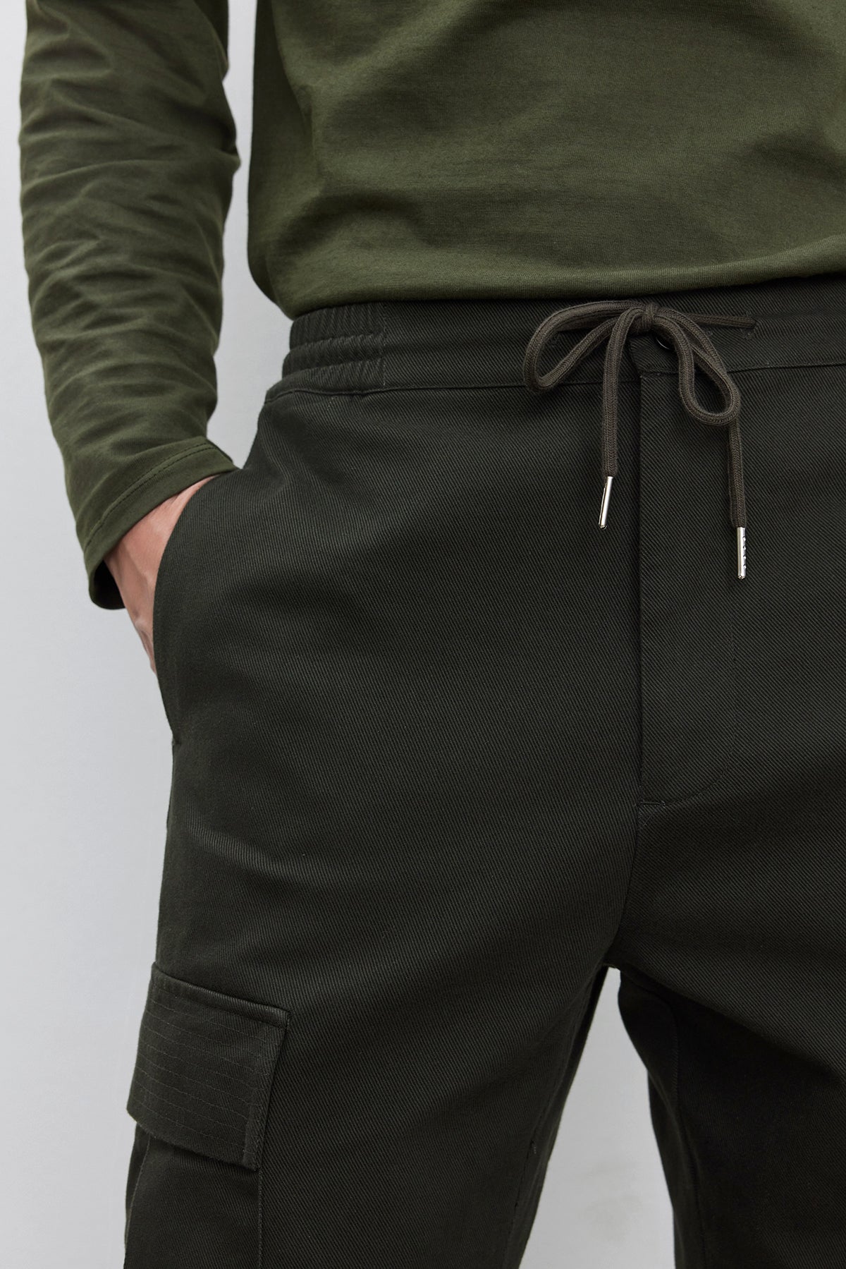   Person wearing Velvet by Graham & Spencer's dark green BRYCE PANT made of mid-weight cotton lyocell twill with an elasticized drawstring waist and a dark green long-sleeve shirt, with one hand in a pocket. 