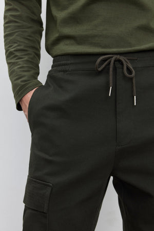 Person wearing Velvet by Graham & Spencer's dark green BRYCE PANT made of mid-weight cotton lyocell twill with an elasticized drawstring waist and a dark green long-sleeve shirt, with one hand in a pocket.