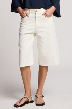 A person wears Velvet by Graham & Spencer’s OAKLEY DENIM SHORT in off-white with a navy top and tan sandals, standing against a plain background with hands in pockets.