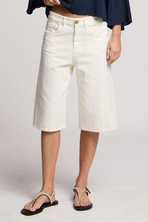 Model wears Velvet by Graham & Spencer OAKLEY DENIM SHORT, paired with a dark blue top and beige sandals. Shown from shoulders down against a plain background.