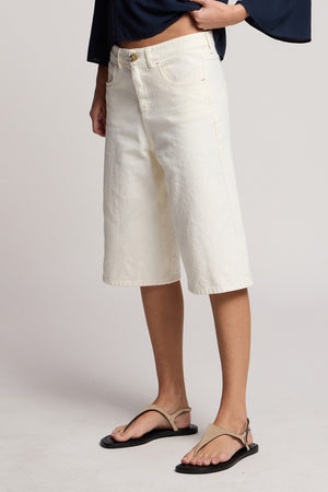 A person wearing Velvet by Graham & Spencer’s OAKLEY DENIM SHORT in off-white, paired with a navy top and beige sandals, stands against a neutral background.