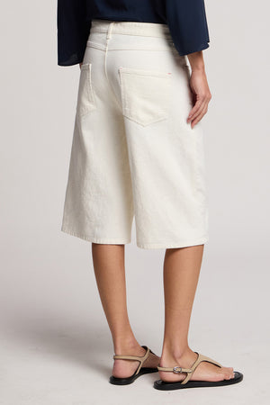 A person shown from the back, wearing Velvet by Graham & Spencer's OAKLEY DENIM SHORT in white, paired with a dark top and beige sandals against a neutral background.