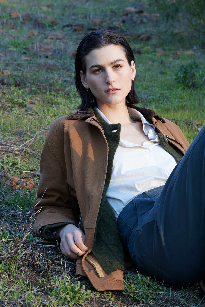 Person with short dark hair wearing the OAKLEY JACKET by Velvet by Jenny Graham, featuring corduroy trims, and a white shirt, lying on grass outdoors.