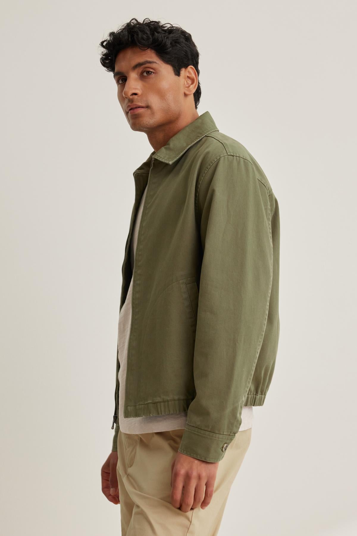   A person stands sideways in an olive COLLINS JACKET by Velvet by Graham & Spencer, paired with a white shirt and beige pants against a neutral background. 