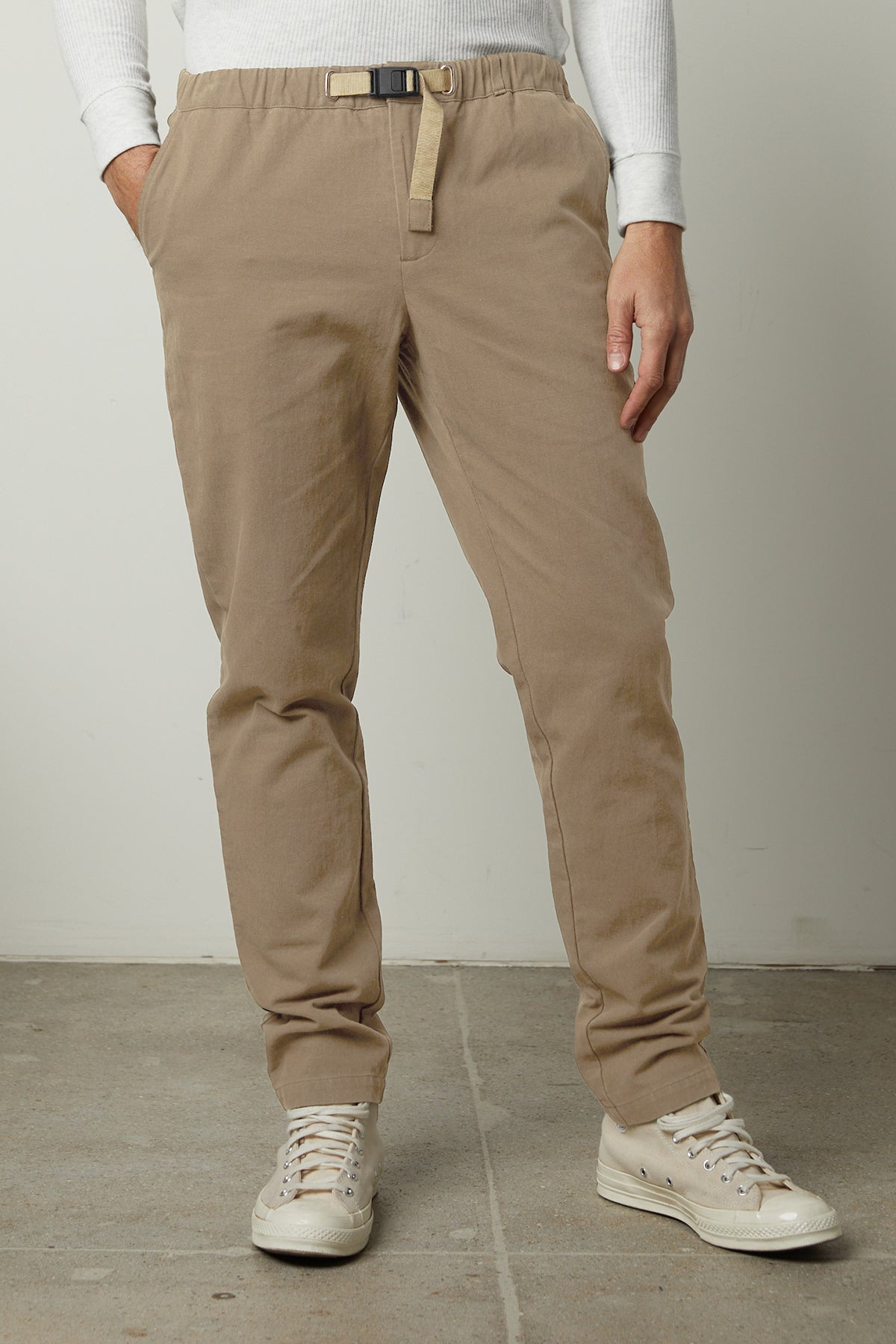   A man wearing the MORAN PANT by Velvet by Graham & Spencer in beige, which features a built-in woven belt, paired with a white long-sleeve shirt and white sneakers. His hands are in his pockets as he stands on a concrete floor. 