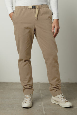 A man wearing the MORAN PANT by Velvet by Graham & Spencer in beige, which features a built-in woven belt, paired with a white long-sleeve shirt and white sneakers. His hands are in his pockets as he stands on a concrete floor.