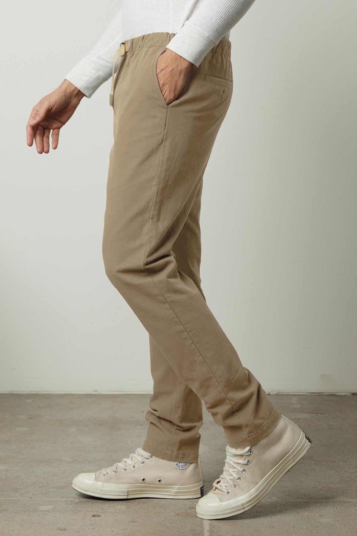   A person wearing the MORAN PANT by Velvet by Graham & Spencer—a beige pant with an elastic waist—and white sneakers stands against a neutral background, with only the lower half of their body visible. 