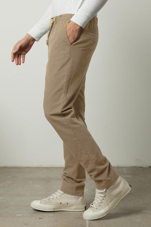 A person wearing the MORAN PANT by Velvet by Graham & Spencer—a beige pant with an elastic waist—and white sneakers stands against a neutral background, with only the lower half of their body visible.