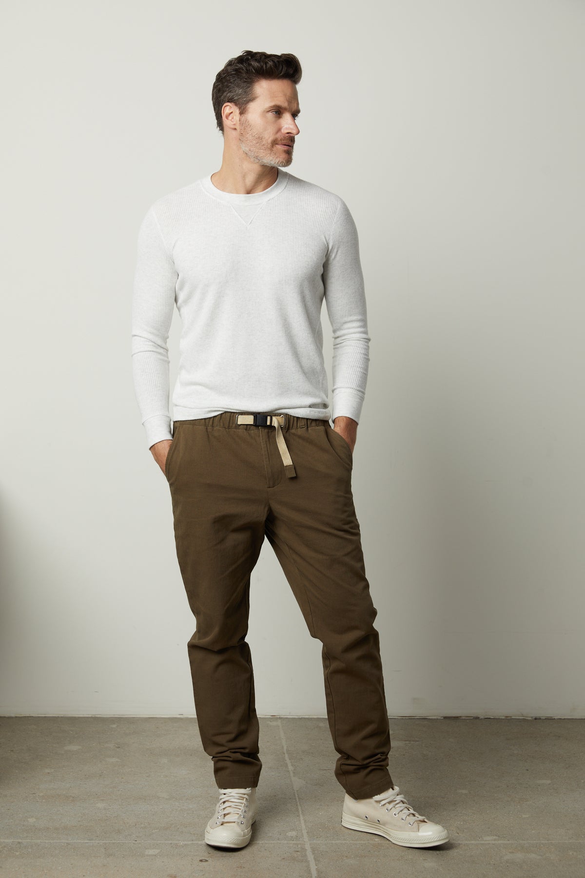 A man stands against a plain background, wearing a white long-sleeve shirt, MORAN PANT by Velvet by Graham & Spencer made of cotton twill fabric with an elastic waist, and light-colored sneakers. His hands are in his pockets.-26768108781761