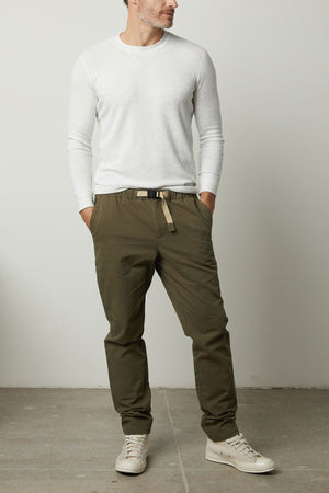 A man wears the Velvet by Graham & Spencer MORAN PANT—olive green cotton twill joggers with a belt—paired with a white long-sleeve shirt and beige sneakers, standing against a plain white wall for a modern, versatile look.