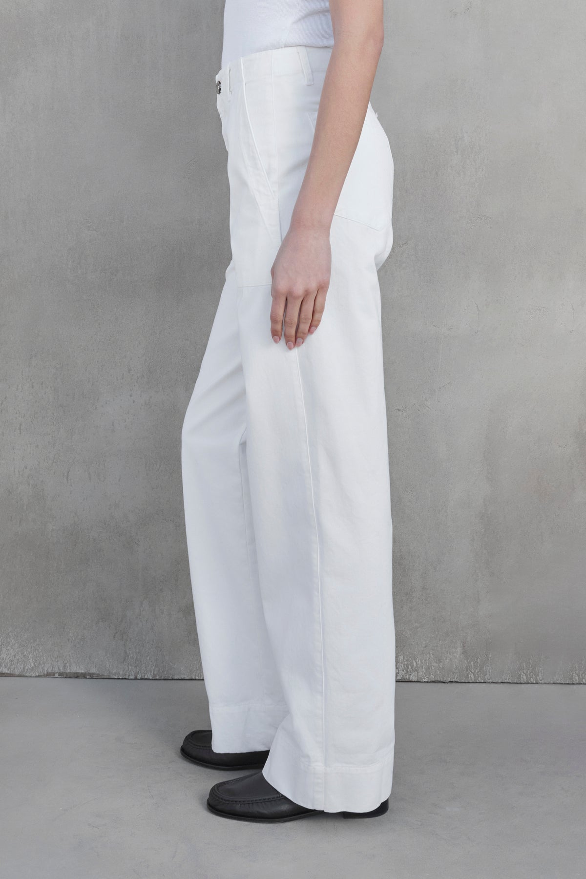   Side view of a person wearing the VENTURA PANT by Velvet by Jenny Graham, styled with a white top and black shoes, standing against a gray textured wall. 