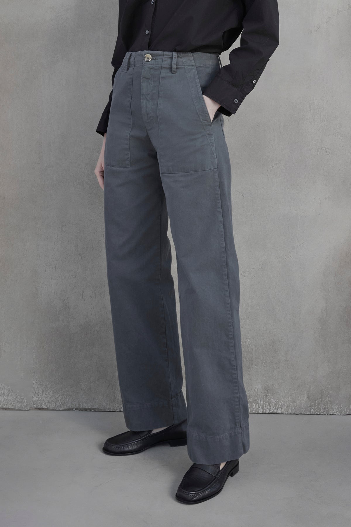   Person wearing a black long-sleeve shirt tucked into the VENTURA PANT by Velvet by Jenny Graham, which are high-waisted, wide-leg grey cotton twill pants, and black shoes standing against a grey wall. 