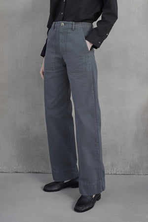 Person wearing a black long-sleeve shirt tucked into the VENTURA PANT by Velvet by Jenny Graham, which are high-waisted, wide-leg grey cotton twill pants, and black shoes standing against a grey wall.