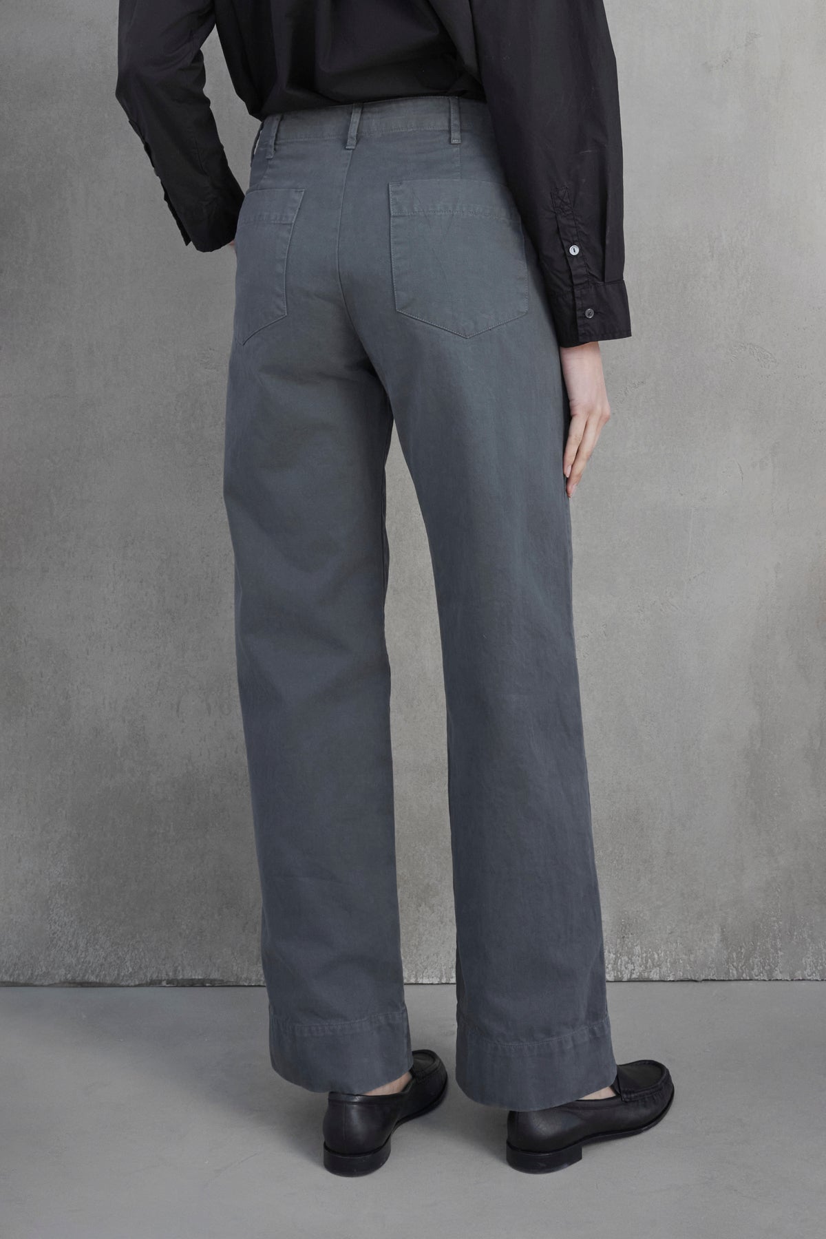   A person wearing the VENTURA PANT in gray cotton twill by Velvet by Jenny Graham and a black long-sleeve shirt is standing with their back to the camera, against a gray concrete wall, exuding a utilitarian vibe. 