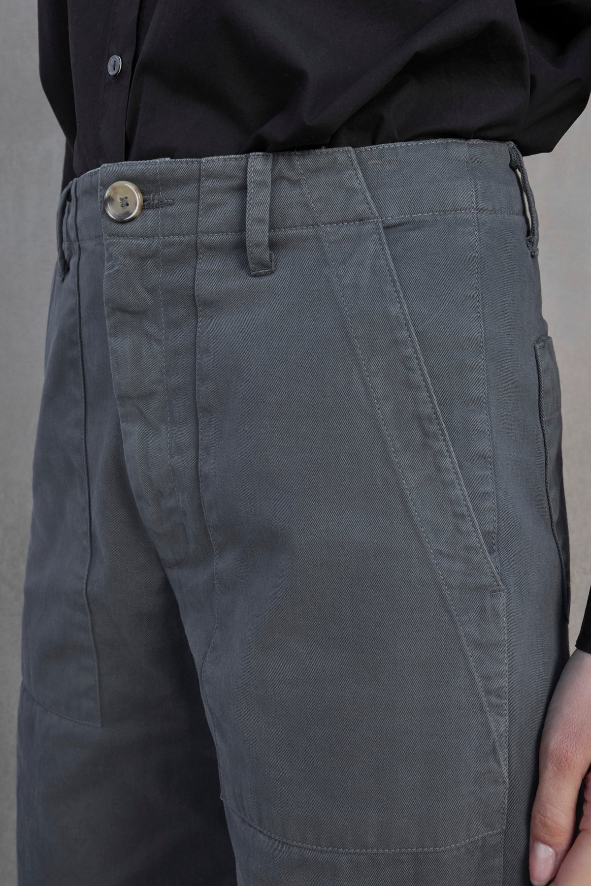   Close-up of a person wearing VENTURA PANT from Velvet by Jenny Graham, featuring grey, cotton twill fabric with a buttoned waist and front pockets, paired with a black shirt. 