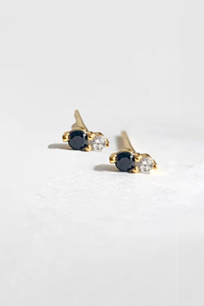 Close-up of Twin Stud Onyx by Phyllis and Rosie earrings with paired black and clear stones on gold posts on light gray