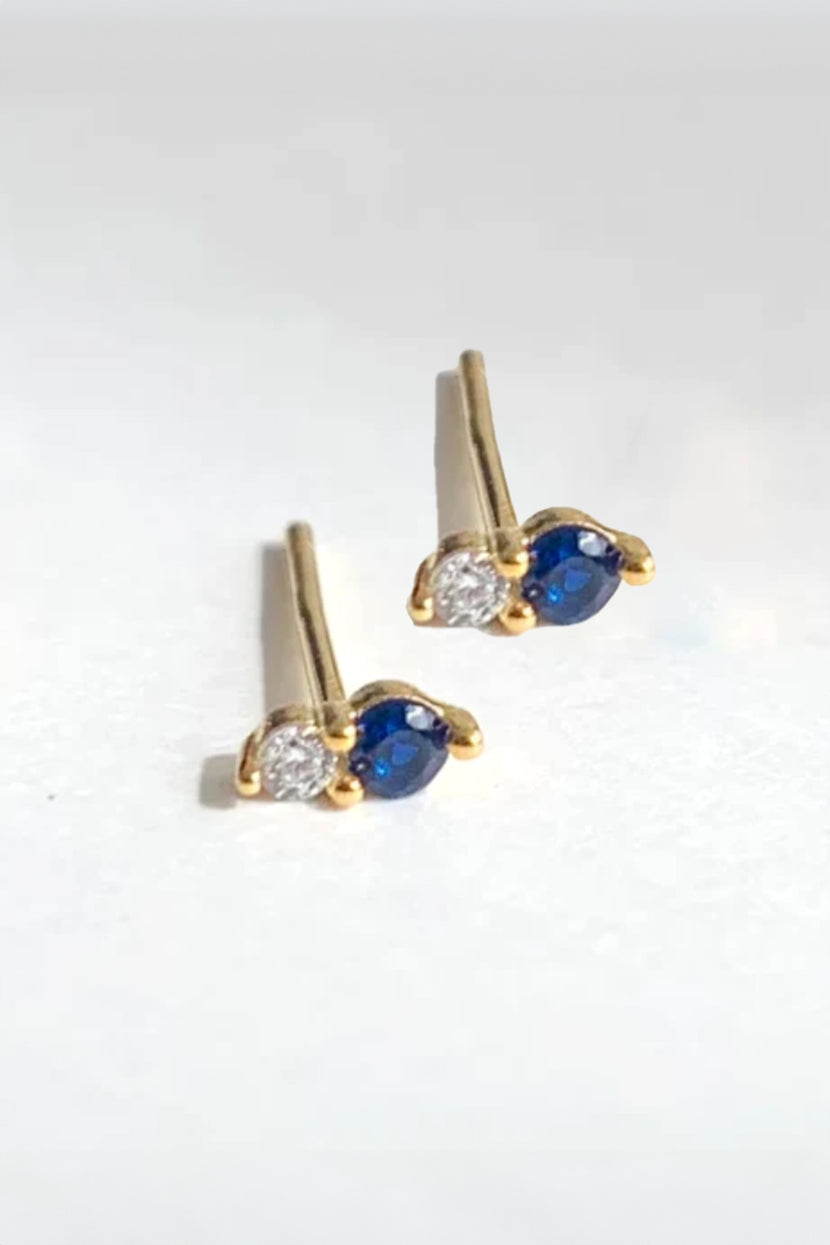 A pair of chic Twin Stud Sapphire earrings by Phyllis+Rosie, featuring one blue and one clear gemstone on each earring, placed on a white surface.