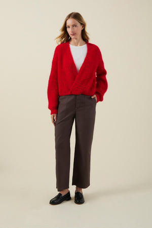 A woman stands against a plain light background wearing the vibrant red RANYA ALPACA CARDIGAN by Velvet by Graham & Spencer, styled with a white shirt, brown wide-leg pants, and black loafers.