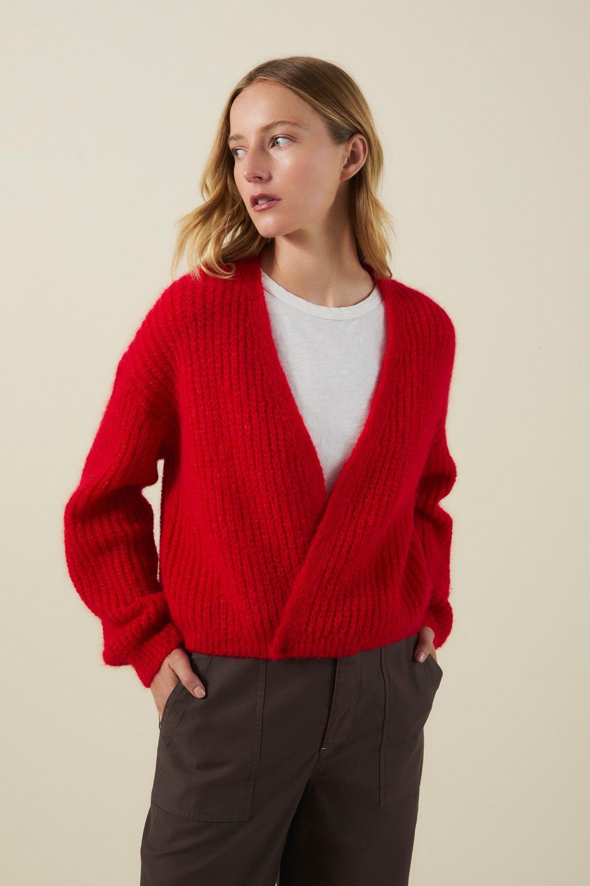 Wearing the RANYA ALPACA CARDIGAN by Velvet by Graham & Spencer—a bright red, chunky knit, wrap-style cardigan—layered over a white shirt and dark pants, this person stands with hands in pockets against a plain background.