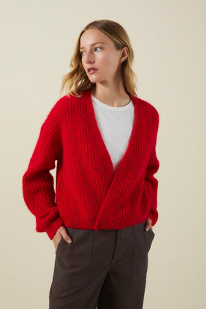 Wearing the RANYA ALPACA CARDIGAN by Velvet by Graham & Spencer—a bright red, chunky knit, wrap-style cardigan—layered over a white shirt and dark pants, this person stands with hands in pockets against a plain background.