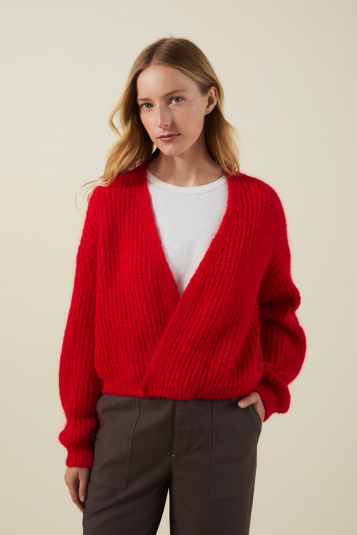 A woman stands against a plain light background, layering the Velvet by Graham & Spencer RANYA ALPACA CARDIGAN—a bright red, alpaca blend wrap-style piece—over a white shirt and dark trousers for the perfect cozy look.-41976490557633