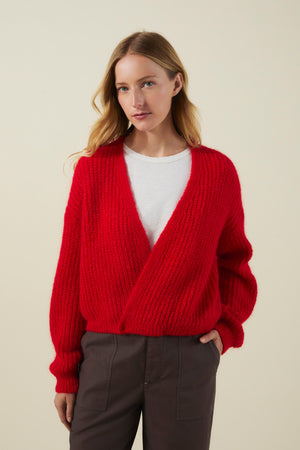 A woman stands against a plain light background, layering the Velvet by Graham & Spencer RANYA ALPACA CARDIGAN—a bright red, alpaca blend wrap-style piece—over a white shirt and dark trousers for the perfect cozy look.