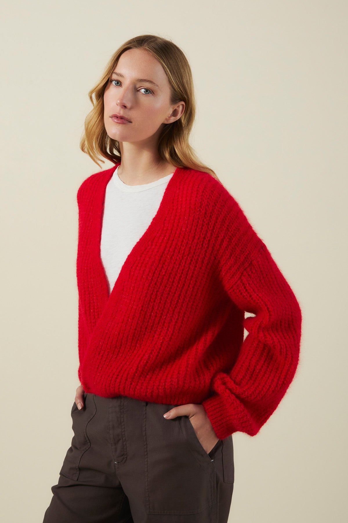 A woman wears the RANYA ALPACA CARDIGAN by Velvet by Graham & Spencer—a red, V-neck chunky knit—layered over a white shirt and paired with dark pants, standing against a plain beige background.-41976490590401