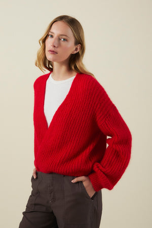 A woman wears the RANYA ALPACA CARDIGAN by Velvet by Graham & Spencer—a red, V-neck chunky knit—layered over a white shirt and paired with dark pants, standing against a plain beige background.