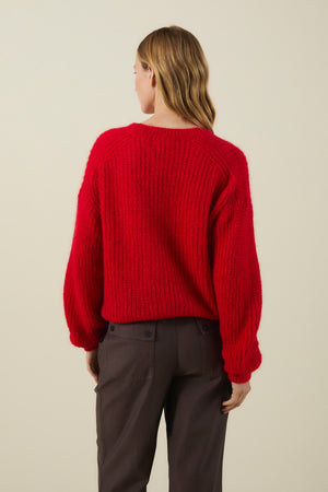 An individual with long hair is seen from behind, standing against a plain background and wearing the Velvet by Graham & Spencer RANYA ALPACA CARDIGAN in a vibrant red hue paired with dark pants. The cardigan’s cozy alpaca blend fabric and elegant wrap-style silhouette bring both warmth and sophistication to the look.