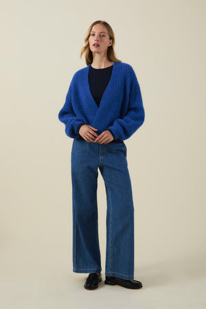 A person stands against a plain background wearing the RANYA ALPACA CARDIGAN in blue by Velvet by Graham & Spencer, paired with a black shirt, wide-leg blue jeans, and black shoes.