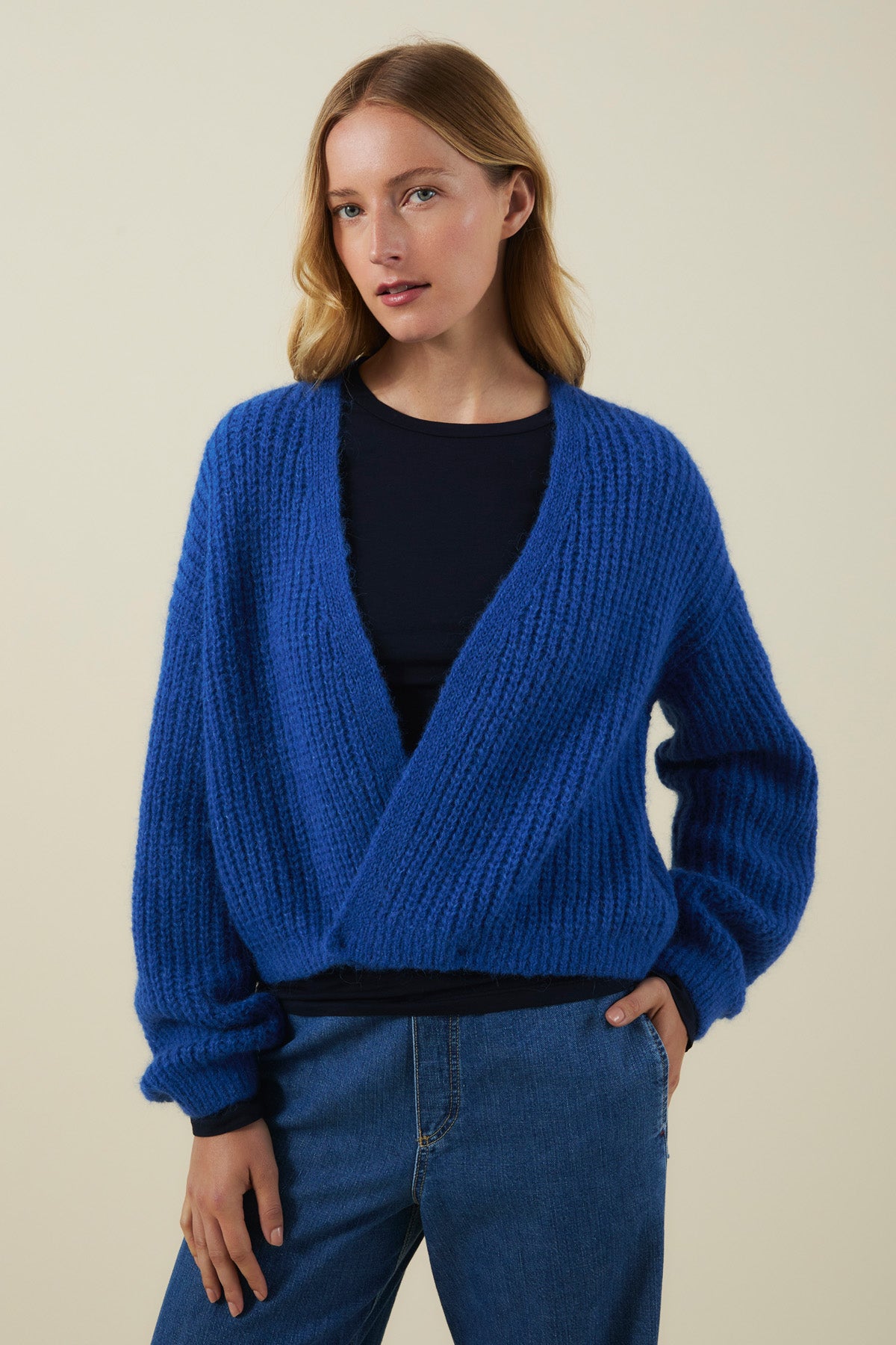 Wearing the RANYA ALPACA CARDIGAN by Velvet by Graham & Spencer in royal blue over a black top and blue jeans, a person stands against a plain beige background.-41976490754241