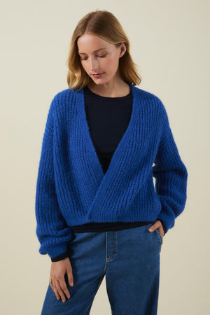 A woman wears the RANYA ALPACA CARDIGAN by Velvet by Graham & Spencer—a textured blue alpaca blend piece—layered over a black top and blue jeans. She is standing against a plain beige background, looking down with her hands resting by her sides.